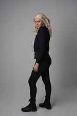  full length portrait of a blonde girl wearing  modern black jacket and pants, standing pose  facing away from the camera on grey studio background.