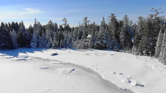 Scenic aerial flyby of remote Winter shoreline with snow covered evergreen trees and secluded cottages.
