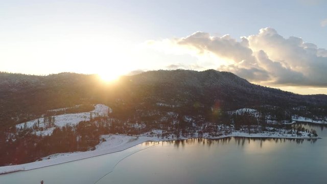 Dazzling aerial shot turning over a lake in winter with bright sun and lens flare. Snowy ground and hills.