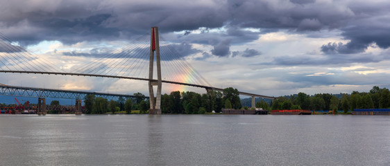 Obraz premium Beautiful view of Skytrain Bridge going over Fraser River during a cloudy evening with rainbow in the background. Taken in New Westminster, Vancouver, BC, Canada.