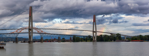 Beautiful view of Skytrain Bridge going over Fraser River during a cloudy evening with rainbow in the background. Taken in New Westminster, Vancouver, BC, Canada.