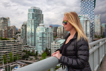 Fototapeta na wymiar Blond young girl is looking at the scenery from Granville Bridge in the modern city during a cloudy evening. Taken in False Creek, Downtown Vancouver, BC, Canada.