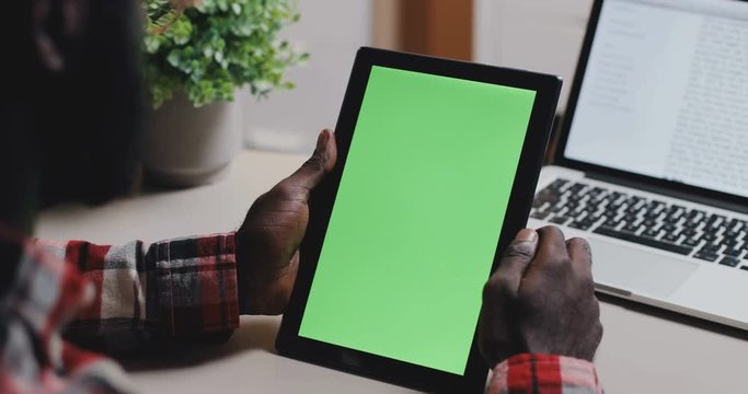 View over the shoulder on the African American man's hands holding black tablet device with chroma key screen and scrolling, tapping and zooming at the table with laptop. Close up.