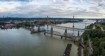 Fototapeta na wymiar Aerial panoramic view of Pattullo and Skytrain Bridge going across Fraser River duing a cloudy evening before sunset. Taken in New Westminster, Vancouver, BC, Canada.