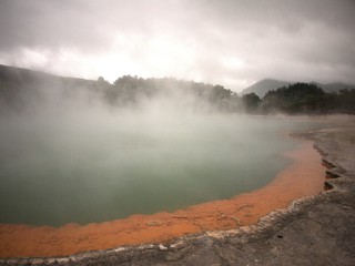 Volcanic rock pool with layers of gray, green, and orange rock with steam rising from water and trees and hills in background under grey, cloudy sky
