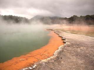 Volcanic rock pool with layers of gray, green, and orange rock with steam rising from water and trees and hills in background under grey, cloudy sky