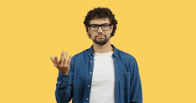 Caucasian man in glasses and jeans shirt doing gesture like so-so with a hand while standing on the yellow screen.