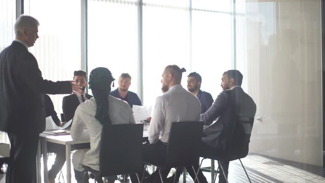 Mature grey-haired financial director reporting to his multiracial top management co-workers of income increase for this year, showing graphs with results of successful teamwork