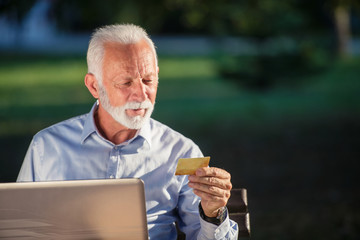 Old male sitting on bench and surfing net on laptop in park, shopping online concept