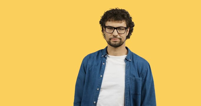 Portrait shot of the Caucasian man in glasses and jeans shirt doing gesture like whatever with a hand while standing on the yellow screen background.
