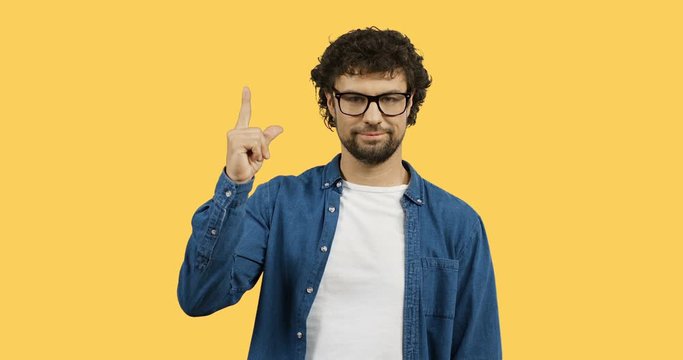 Young handsome man in the glasses and jeans shirt pointing up with his finger in the sky. Yellow back screen.