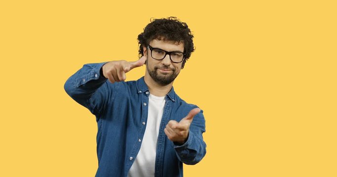 Young handsome man in glasses standing in front of the camera and doing gesture like yeah you with both hands. Yellow screen on the background.