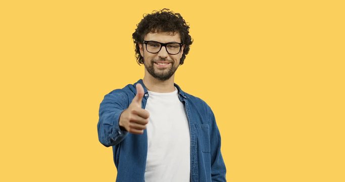 Attractive cheerful man in glasses with curly hair giving his thumb up while standing on the yellow screen background.