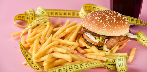 burger and french fries and a sweet drink. Sewing Tape Measures on the food. concept of losing weight and unhealthy diet, getting fat because of junk food. takeaway lunch, pink background