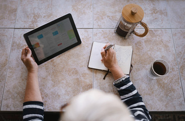 Top view of mature woman making note in her notebook or journal while reading off an article on a...