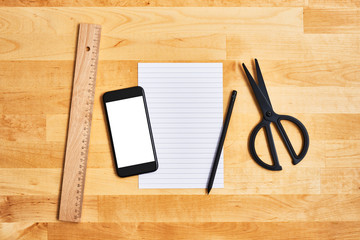 An empty sheet of paper with black smartphone, ruler, pencil and scissors on yellow wooden table. School concept. Top view. Copy space for text.