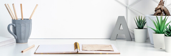 White desk next to an empty wall with space for text. Copy space. Green succulents as a decoration. Opened notebook, office stationery, gray mug, bonsai and graphic element on the wall. Panorama