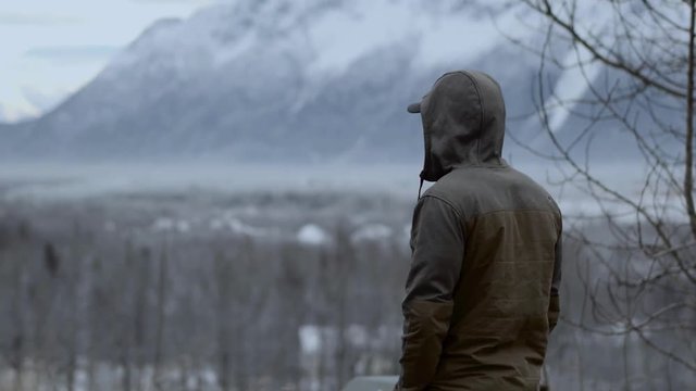 Man in winter jacket looking into the distance, mountains and snow covered landscape in background 4k