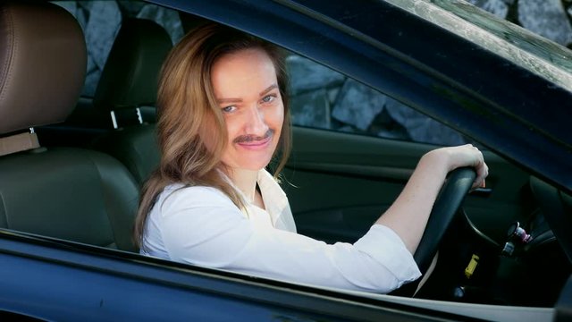 a portrait of an androgynous feminist with a painted mustache on her face sitting behind the wheel of a car. woman looks into the camera from the car window.