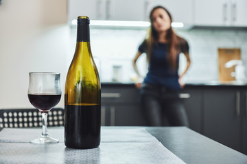 You don’t get over an addiction by stopping using. You recovery by creating a new life where it’s easier to not use. A bottle and glass of wine on the table. Addicted woman standing in the background - obrazy, fototapety, plakaty