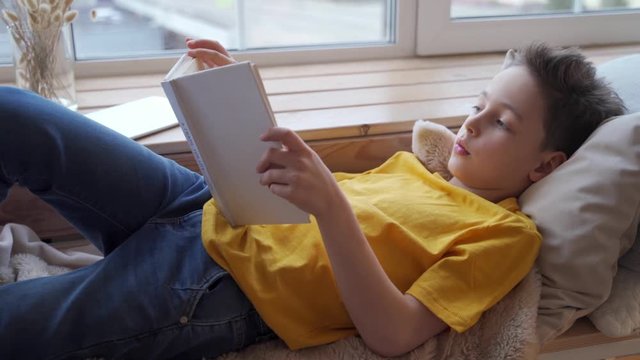 Cute little boy lying on windowsill and reading book