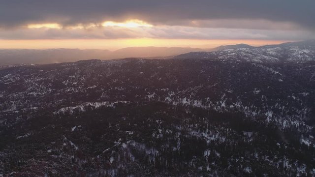 Aerial shot turning over snowy forest at sunset. Beautiful clouds with sun glow behind near sunset. Mountains in California Sierras.