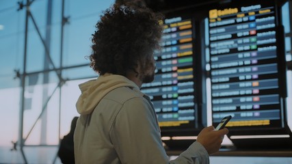 Young middle aged traveler man with curly hair and backpack stands in airport departure terminal compares data from email and flight information display system to look for boarding gate or delay.