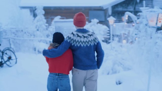 Young couple in winter knit clothing walks together towards hotel or apres ski restaurant on cold winter day, hugging and cuddling during romantic dreamy vacation. Perfect winterland destination
