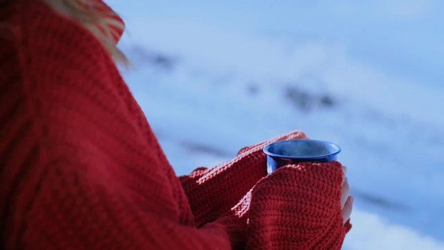 Young millennial hipster traveler on winter vacation or camping trip, wears red knit sweater and sips coffee or tea from mug or cup on cold freezing day, beverage evaporates. Outdoor lifestyle