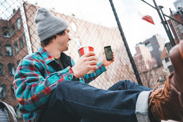 Young trendy man sitting on the street sidewalk resting with cup of hot coffee and using smartphone for video link. Casual man calling on video link his friends