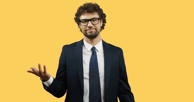 Good looking Caucasian businessman in glasses, suit and tie standing on the yellow wall screen and doing gesture so-so or fifty-fifty with a hand.