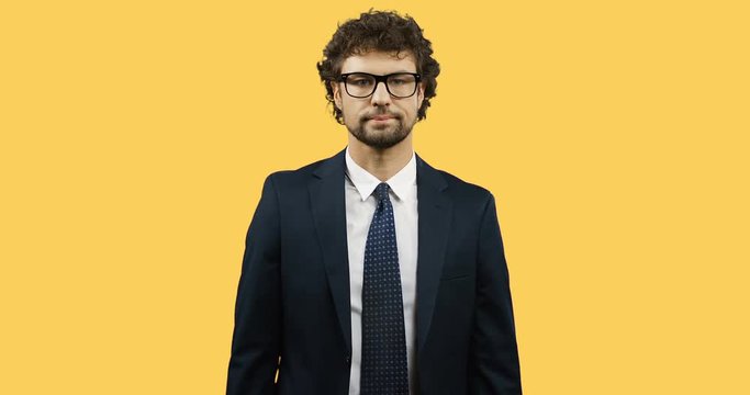 Attractive Caucasian businessman in glasses, suit and tie standing on the yellow back screen and waving his hand like whatever or let it be.