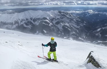 Fotobehang Lover of ski in low Tatras waiting for best weather. A view on freeride. A man skier standing on the top of mountain and choosing best track. Yellow helmet, black backpack © Fauren