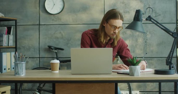 Camera zooming in on the young good looking stylish guy in glasses and with long hair working at the laptop computer and then writing something down in the notebook at his urban office. Portrait shot.