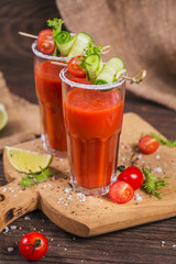 Two glasses of fresh organic tomato juice decorated with raw tomatoes, cucumber and leaves on a rustic wooden cutting board
