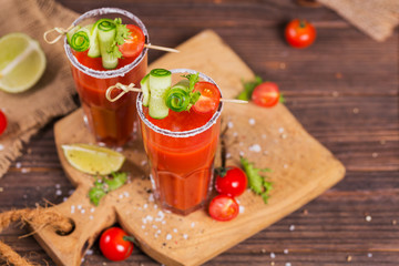 Two glasses of tomato juice decorated with fresh tomatoes, cucumber and leaves on a wooden...