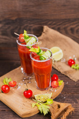 Two glasses of tomato juice decorated with fresh tomatoes, cucumber and leaves on a wooden background