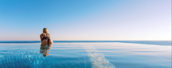 concept of vacation by the sea in the pool. girl in a luxurious swimsuit on the edge of a panoramic pool against the sea - obrazy, fototapety, plakaty
