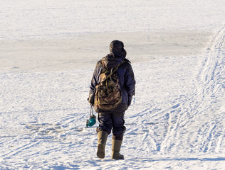 Fototapeta na wymiar A man goes to the river for fishing in the winter. In the hands of tackle for fishing. Backpack on the back. The river is frozen. Footprints in the snow.