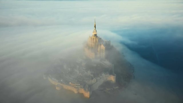 Drone flying very close near ethereal Mont Saint Michel island castle covered by atmospheric sunrise mist in Normandy.