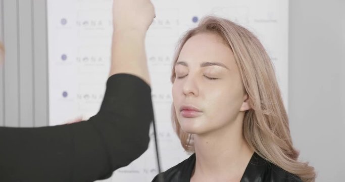 Blonde makeup artist using an airbrush to cover skin imperfections. Beautiful blone girl sits in a chair at a beauty salon.