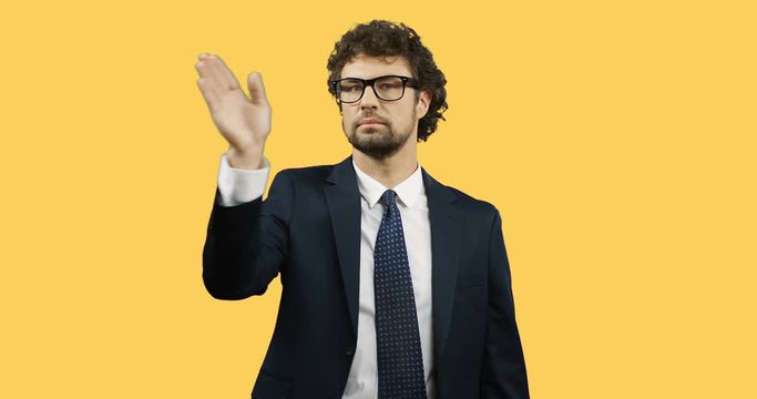 Good looking man in the glasses, suit and tie standing on the yellow wall background, scrolling and taping in the air.