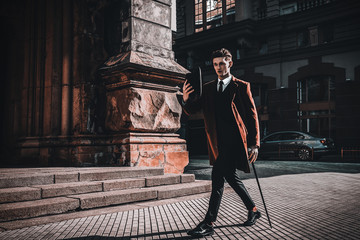 Fashion portrait of young man on red coat, white shirt, black suit and cane walking on streets of...