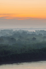 Mystical view on riverbank of large island with forest under haze at early morning. Mist among layers from tree silhouettes under predawn sky. Morning calm atmospheric landscape of majestic nature.