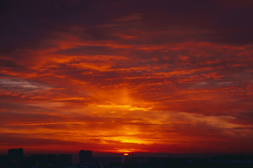 Naklejka premium Cityscape with vivid fiery dawn. Amazing warm dramatic cloudy sky above dark silhouettes of city buildings. Orange sunlight. Atmospheric background of sunrise in overcast weather. Copy space.