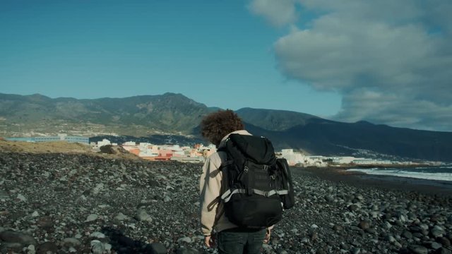 Mysterious, romantic and dramatic looking middle aged hipster man wit backpack during hiking, camping or backpacking trip walks on windy epic beach with mountains in background, looks back at camera