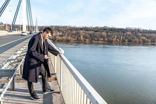 Depressed and anxious man standing on the bridge with suicidal thoughts disappointed in people looking down to jump in the water and end life with sending farewell message or suicide note by phone 