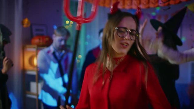 Halloween Costume Party: Beautiful She Devil with Trident and Horns Dances Charmingly. In the Background Zombie, Death, Witch and Mummy Have Fun in a Monster Party Decorated Room