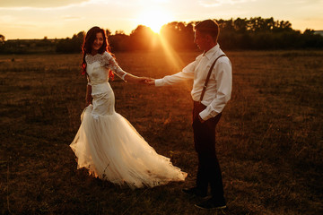 Fototapeta na wymiar Newlyweds are running across the field, holding hands. Beautiful girl in a white dress and stylish groom, beautiful sunset.