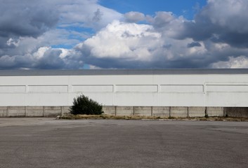 Fototapeta na wymiar White industrial warehouse building with a cement surrounding wall and a large asphalt road under a blue sky with many clouds. Background for copy space